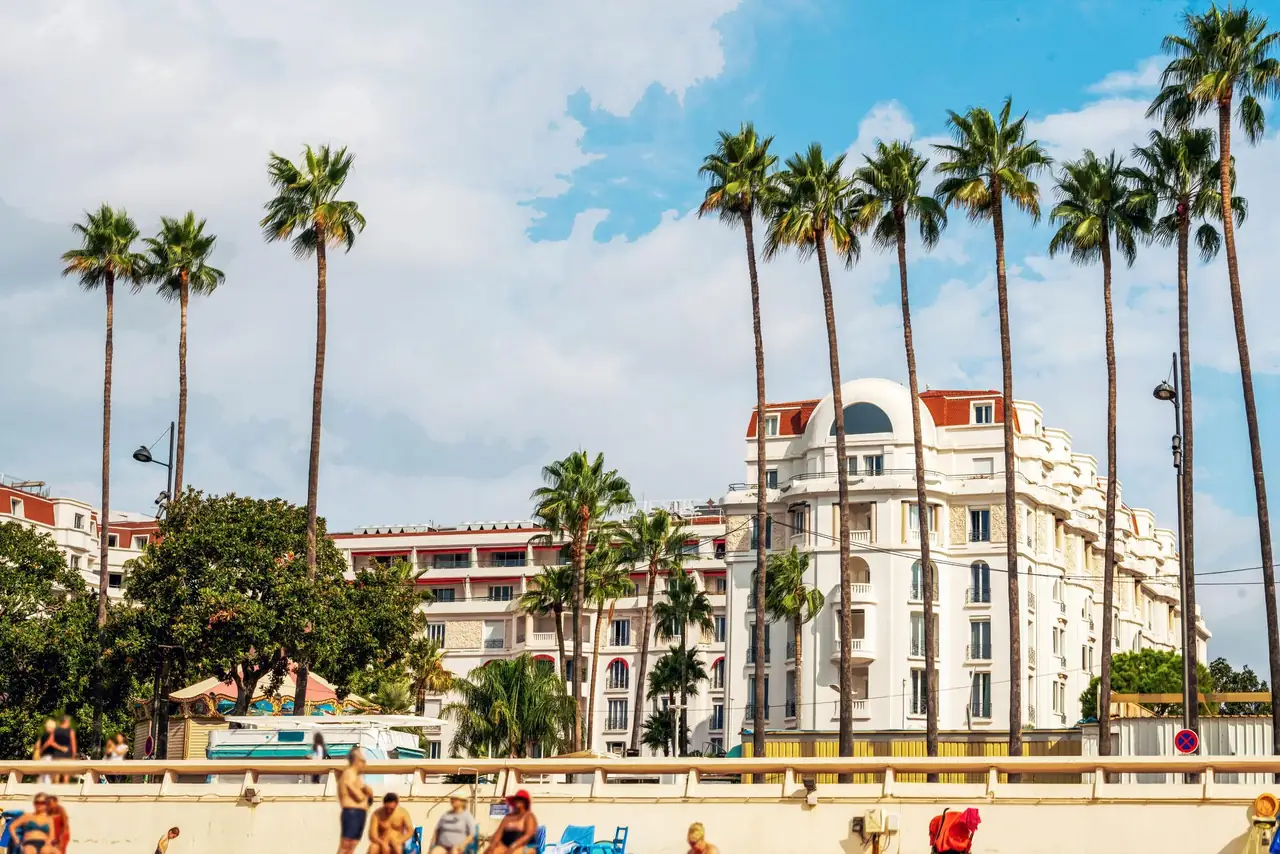 La façade blanche d'un palace de la Croisette à Cannes, encadrée de hauts palmiers, vue depuis la plage